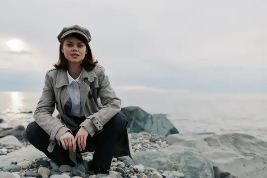 Молодая женщина в кепке и плаще сидит на камнях у моря
