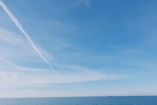 Открытый морской пейзаж с глубокой синей водой, четким горизонтом, далеким судном и следом самолета в небе, безмятежный и обширный