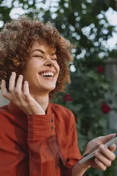 Кудрявая девушка в красной рубашке смеётся, держа телефон на улице