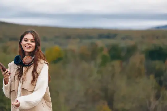 Улыбающаяся девушка в наушниках со смартфоном на фоне осенних холмов