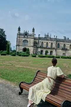 Девушка в светлом платье отдыхает на скамейке у старинного замка