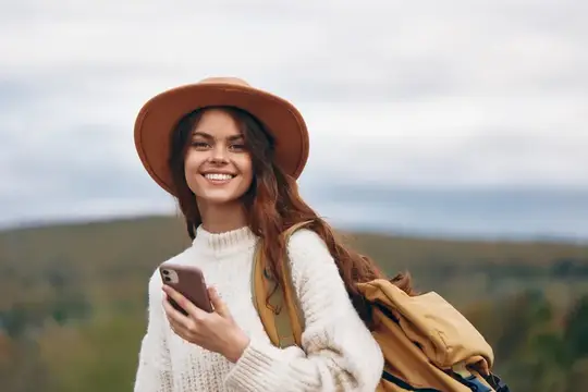 Улыбающаяся девушка в шляпе с рюкзаком держит телефон на фоне холмов