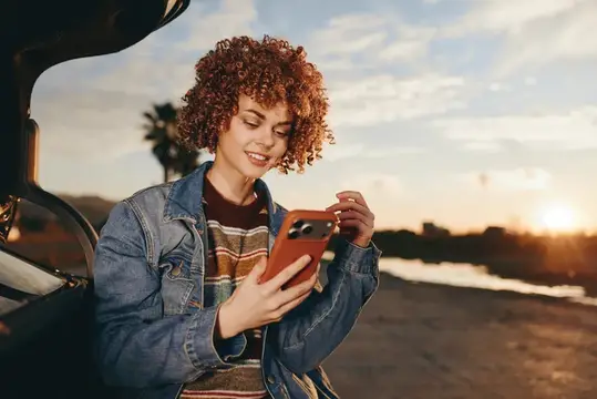 Улыбающаяся женщина с кудрявыми волосами проверяет смартфон, стоя у автомобиля, одета в джинсовую куртку
