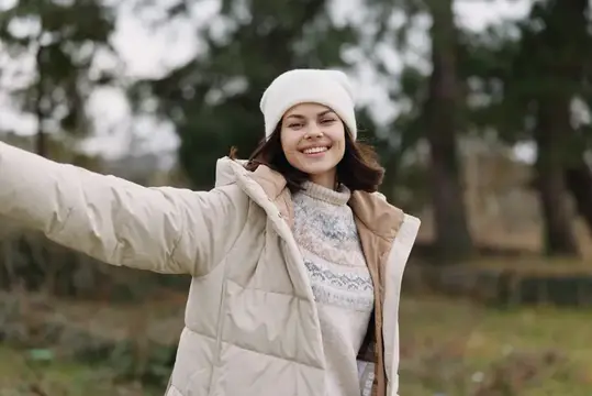 Улыбающаяся девушка в белой шапке и пуховике гуляет по осеннему парку