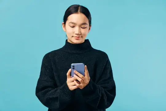 Женщина в уютном черном свитере пользуется смартфоном с легкой улыбкой на светло-голубом фоне, взаимодействие с цифровым устройством, современный образ жизни и технологии на изолированном фоне