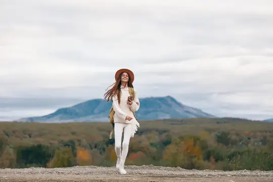 Улыбающаяся девушка в шляпе и белом свитере гуляет на фоне гор
