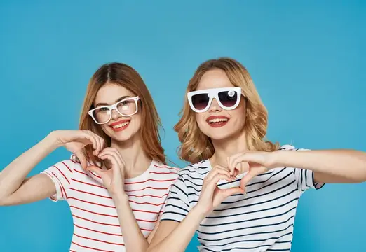 Две улыбающиеся девушки в очках показывают сердечки руками на голубом фоне