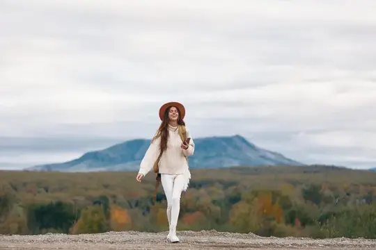 Улыбающаяся девушка в шляпе и свитере гуляет по холму на фоне гор