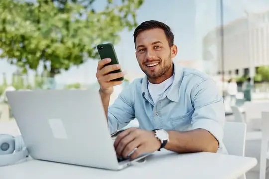 Улыбающийся мужчина с телефоном и ноутбуком за столиком на улице