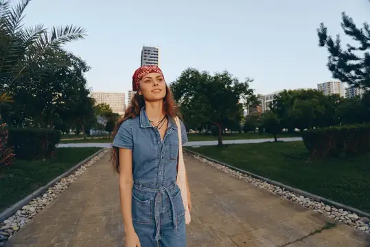 Молодая женщина в джинсовом комбинезоне и красной бандане гуляет по парковой аллее
