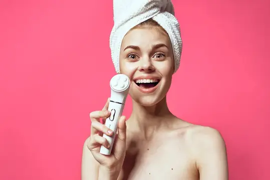Улыбающаяся девушка с полотенцем на голове держит массажёр для лица