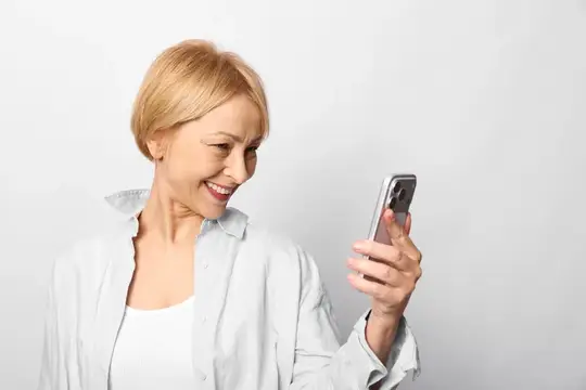 Улыбающаяся блондинка в рубашке смотрит в смартфон на светлом фоне