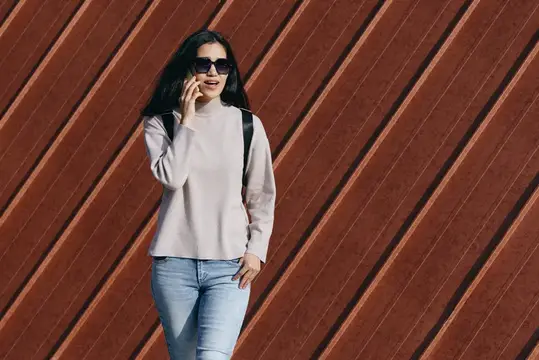 Улыбающаяся брюнетка в солнцезащитных очках говорит по телефону на фоне коричневой стены