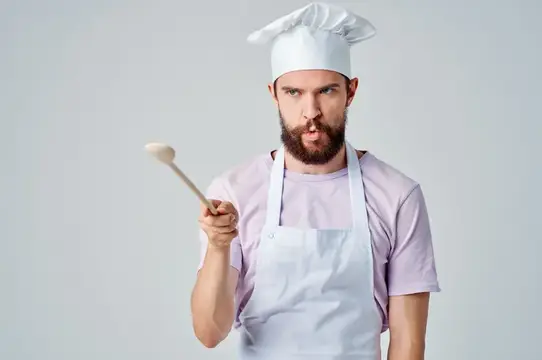 Бородатый мужчина в поварском колпаке пробует соус с половника