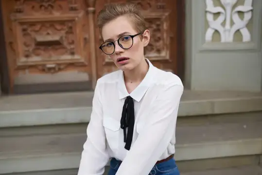 Студентка в джинсах и рубашке сидит на ступеньках возле здания, на лице у неё очки