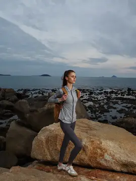 Спортивная женщина в серой спортивной одежде стоит на камнях у моря, демонстрируя задумчивое настроение