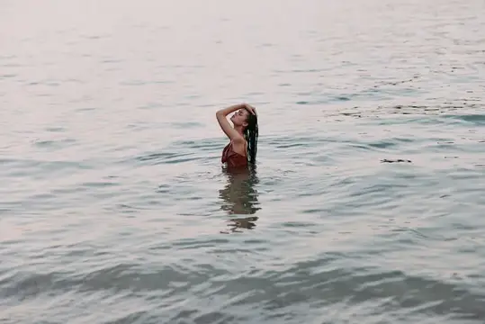 Молодая женщина с темными волосами стоит по грудь в воде и поправляет прическу
