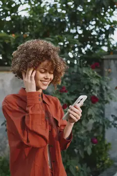 Улыбающаяся кудрявая девушка в терракотовой рубашке смотрит в телефон в саду