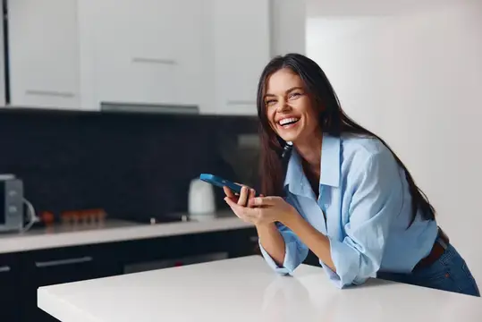 Улыбающаяся брюнетка с телефоном на светлой кухне
