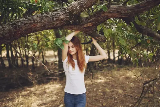 Рыжеволосая девушка в белой футболке стоит под ветвями дерева в саду