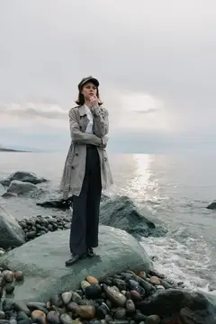 Задумчивая девушка в плаще стоит на камне у моря
