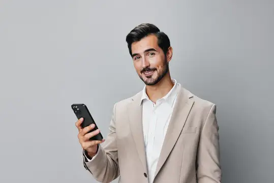 Улыбающийся бородатый мужчина в бежевом пиджаке держит смартфон
