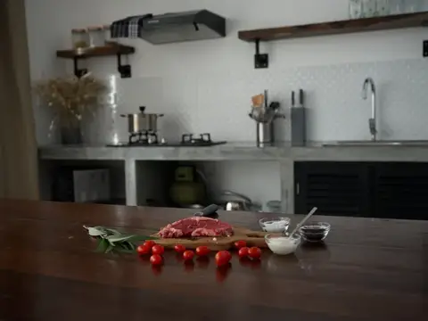 Стильная кухня, на деревянном столе лежит кусок свежего мяса со специями, помидоры и зелень, минималистичный интерьер