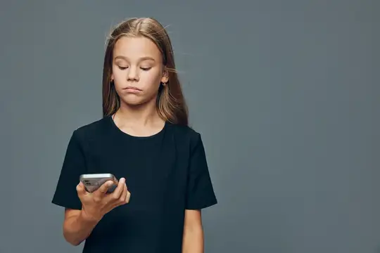 Маленькая девочка держит смартфон, одетая в черную футболку, с нейтральным выражением лица на однотонном фоне