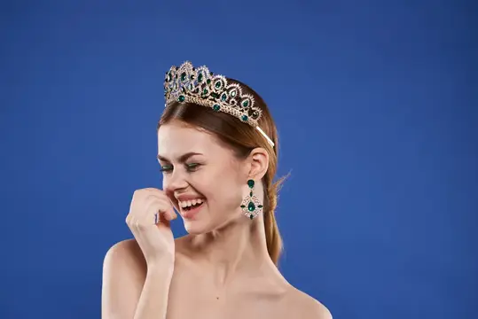 Смеющаяся девушка в короне и серьгах с изумрудами на синем фоне