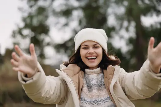 Весёлая женщина в уютной зимней одежде стоит на улице, широко раскинув руки, излучая тепло, радость, открытость и яркое настроение на фоне прохладного уличного пейзажа