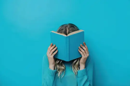 Девушка с распущенными волосами прячет лицо за синей книгой