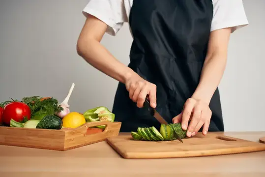 Повар в чёрном фартуке нарезает огурец на доске рядом с овощами