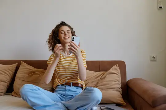 Улыбающаяся кудрявая девушка сидит по-турецки на диване со смартфоном