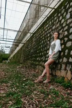 Молодая женщина в белой блузе и шортах прислонилась к каменной стене у теплицы