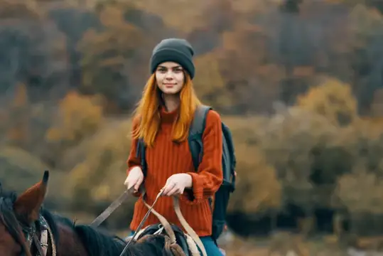 Рыжеволосая девушка в шапке едет верхом на лошади осенью