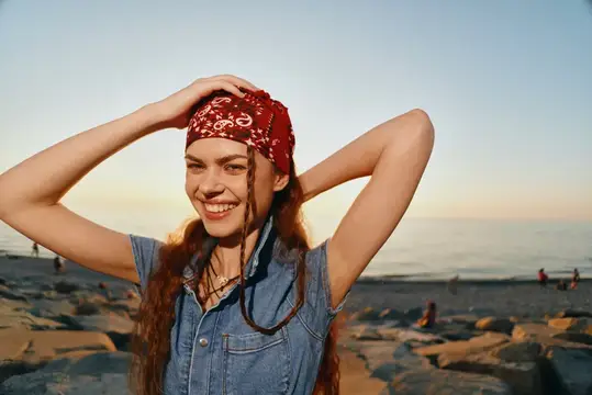 Улыбающаяся девушка в красной бандане поправляет волосы на закатном пляже