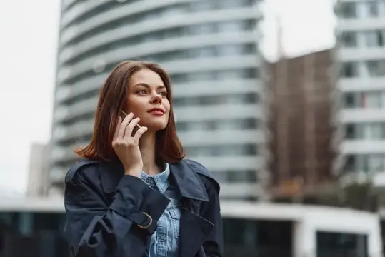 Молодая женщина в плаще говорит по телефону на фоне высоток
