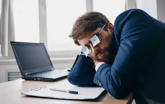 Бизнесмен в очках спит на работе, положив голову на листы бумаги