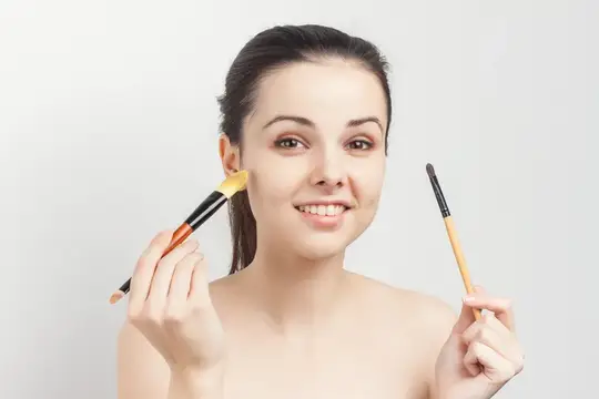 Улыбающаяся девушка с кистями для макияжа у лица на светлом фоне