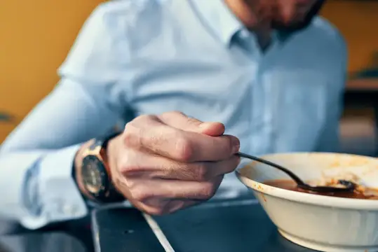 Мужчина в голубой рубашке ест суп ложкой из белой тарелки