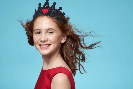 Рыжеволосая девочка в короне с сердечком улыбается на голубом фоне