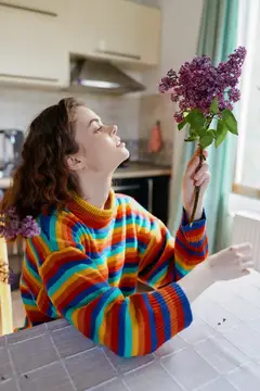 Молодая женщина в разноцветном свитере нюхает ветку сирени на кухне
