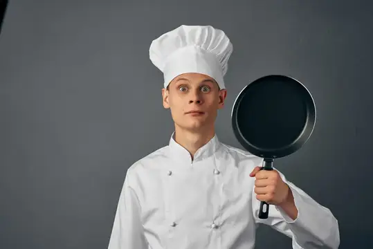 Удивлённый повар в колпаке держит сковороду на сером фоне