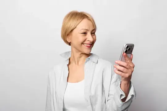 Улыбающаяся блондинка в белой рубашке смотрит в смартфон