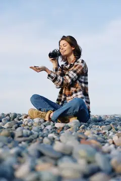 Молодая женщина с фотокамерой сидит на каменистом пляже с закрытыми глазами