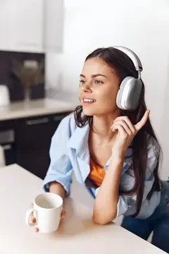 Улыбающаяся темноволосая девушка в наушниках пьёт кофе на кухне