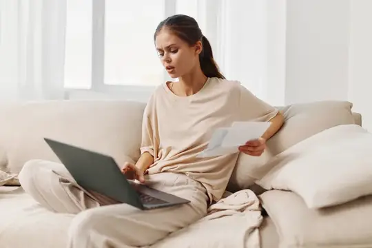 Молодая женщина с документами работает за ноутбуком на диване