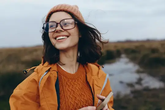 Улыбающаяся девушка в очках и оранжевой куртке на прогулке с телефоном