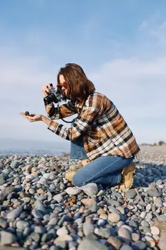 Фотограф стоит на коленях на галечном берегу, выстраивая кадр с помощью портативной камеры