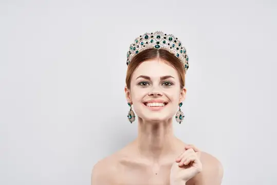 Улыбающаяся рыжеволосая женщина в короне с изумрудами на светлом фоне